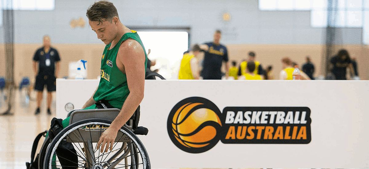 Jordan Bartley in a wheelchair and wearing an Australian training jersey, with a Basketball Australia sign in the background